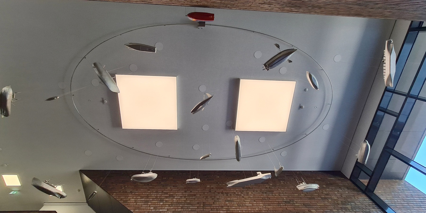 Blick an die Decke des Foyers mit der hängenden Kunstinstallation: Das farbige Boot des Zolls zieht geräuschlos und langsam seine Bahnen zwischen zahlreichen Booten und Schiffen hindurch. © 2020 Staatliches Bau- und Liegenschaftsamt Neubrandenburg