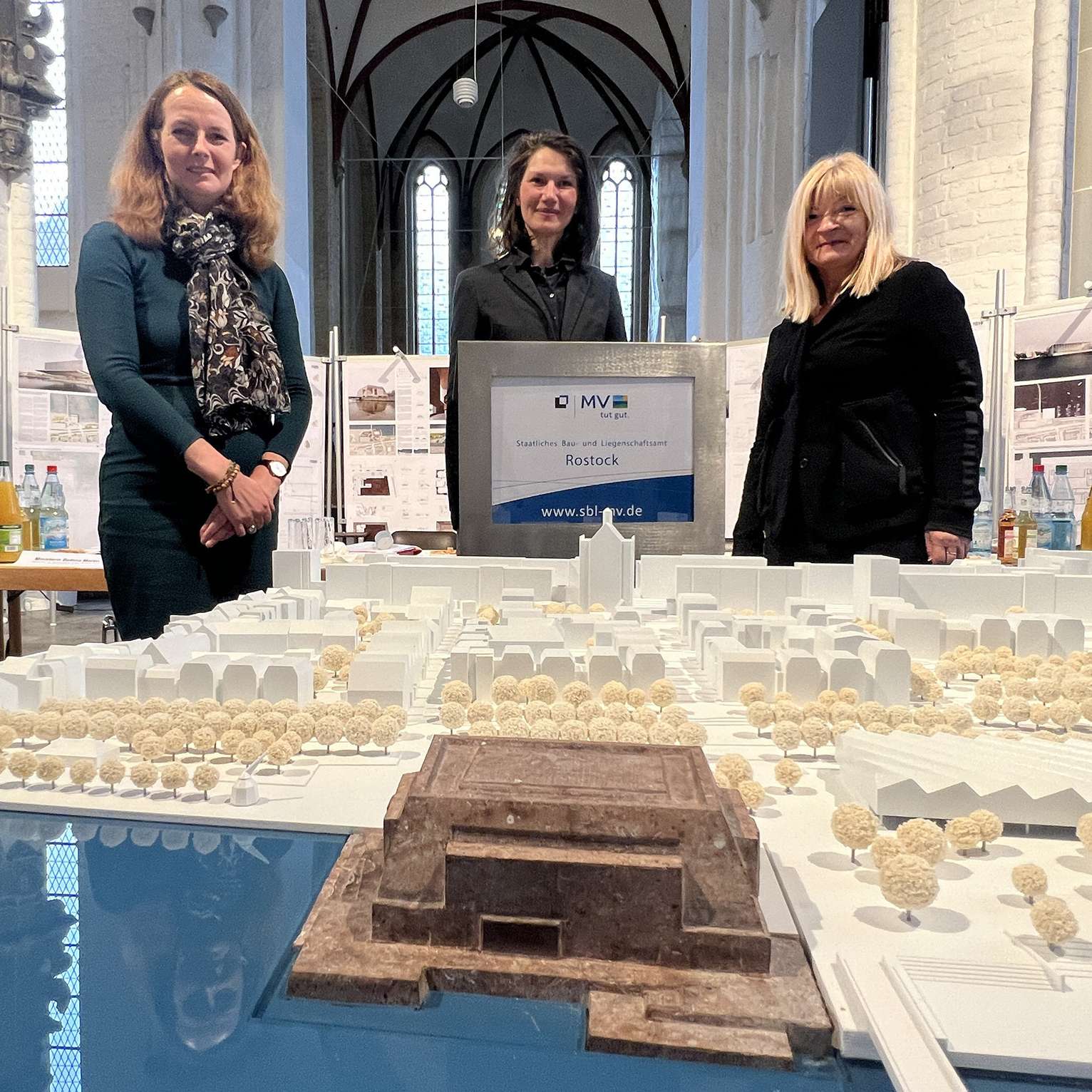 Drei Vertreterinnen des Preisgerichts stehen in der Nikolaikirche hinter dem Modell des Siegerentwurfs. Im Vordergrund ist das Modell zu sehen. © 2022 Christian Hoffmann, FM M-V