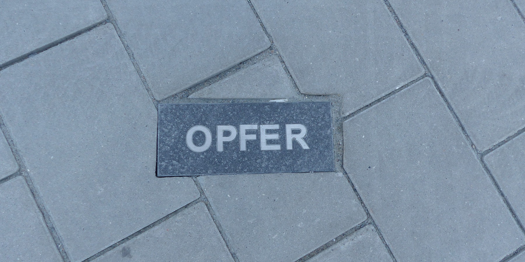 20 eingelassene Granitsteine, davon 8 dunkle mit negativen Begriffen aus der gesellschaftlichen Wirklichkeit (z.B. ANGST, OPFER, GEWALT …) in der Pflaster- bzw. Rasenfläche vor dem Eingang sind Bestandteil der 4-teiligen Kunst am Bau-Installation © 2023 SBL Greifswald