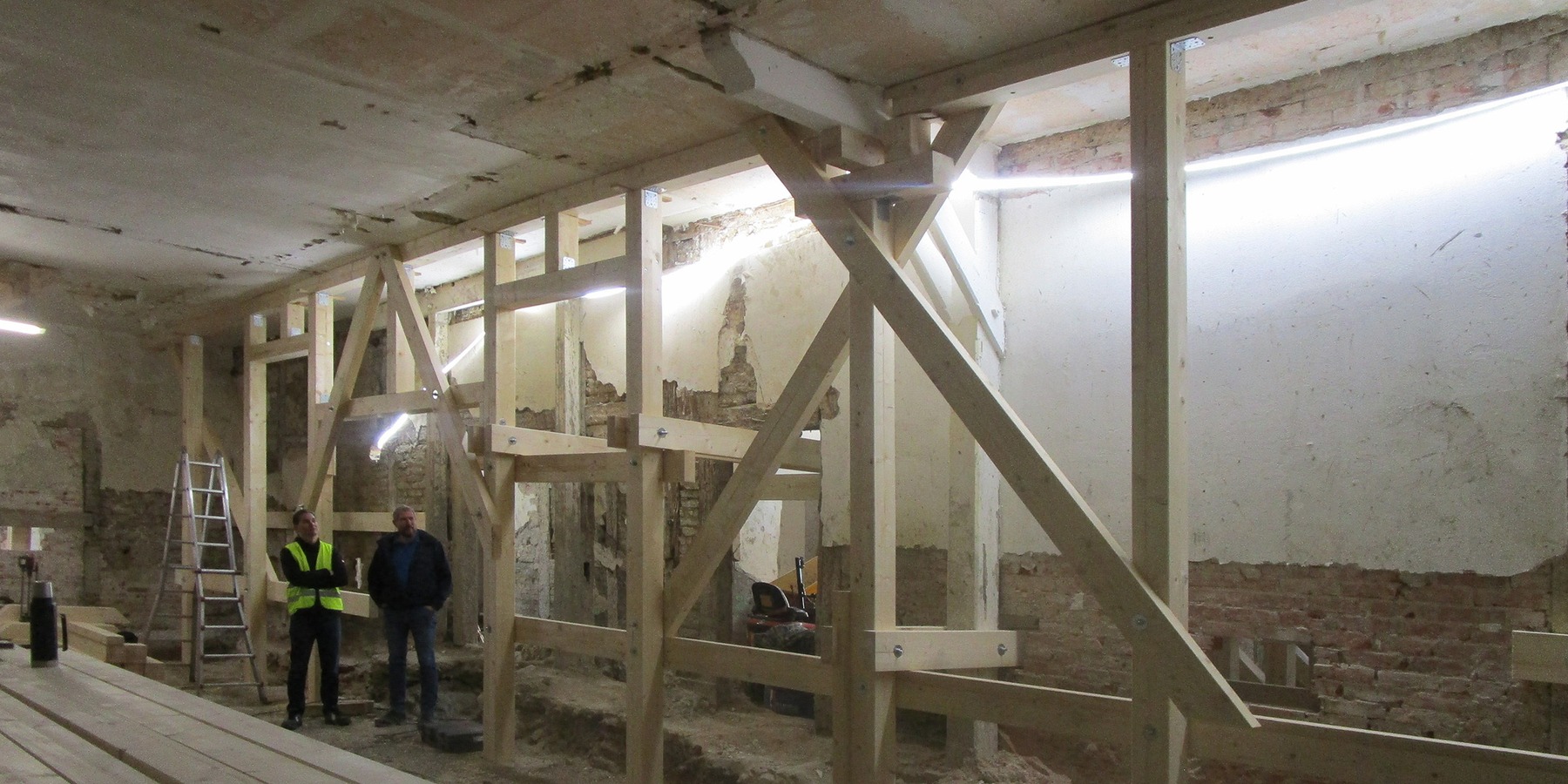 Bautenstand Mai 2026 - vor  Herstellung der neuen Grundplatte und Beginn der Fachwerksanierung mussten provisorische Abfangungen zur Gebäudestabilisierung eingebaut werden - li. Bauleiter Hr. Dahlke (AIU Stralsund, TGA), re. Hr. Lubinski (SBL Greifswald) © 2026 SBL Greifswald